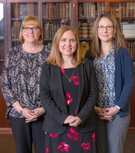 Three women are seen standing in front of a bookcase.